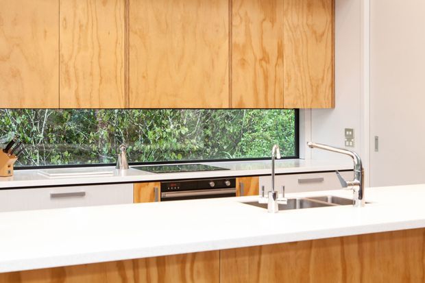 Bright contemporary kitchen featuring a large white island benchtop, integrated stainless-steel sink and tapware, light timber veneer cabinetry, and pale grey lower cabinets. A wide horizontal window provides a leafy outlook and abundant natural light.