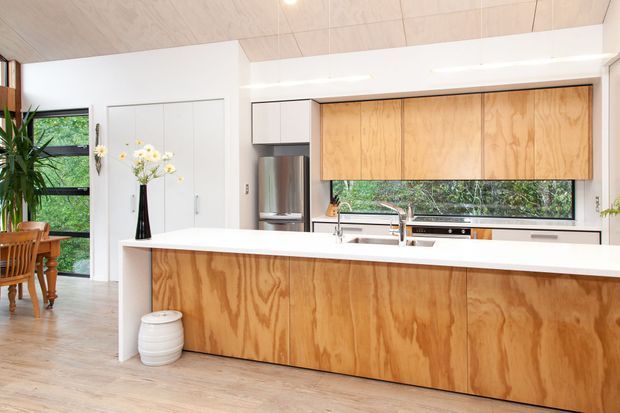 Bright contemporary kitchen with a large white island, natural plywood cabinetry, stainless steel appliances, timber flooring, and a pale timber-lined ceiling. A dining area and garden-facing glazing are visible beyond the kitchen.