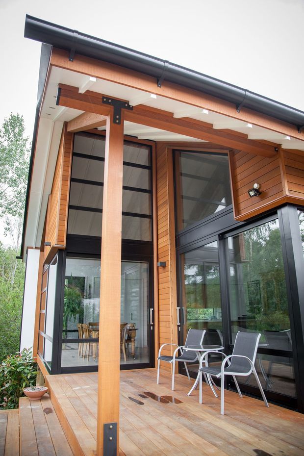 Exterior view of a contemporary timber-clad house featuring a covered timber deck, large black-framed glazed doors and windows, exposed timber posts and beams with dark metal brackets, and outdoor seating. The deck connects to an interior dining area visible through the glazing. A dark guttered roof and surrounding greenery frame the facade.