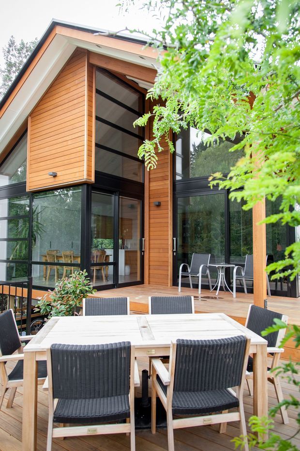 A contemporary timber-clad house with expansive black-framed glazing, viewed from a furnished outdoor timber deck. The two-storey facade features warm horizontal timber boards, a prominent glazed upper-level opening, deep eaves, and an outdoor dining setting surrounded by leafy vegetation.