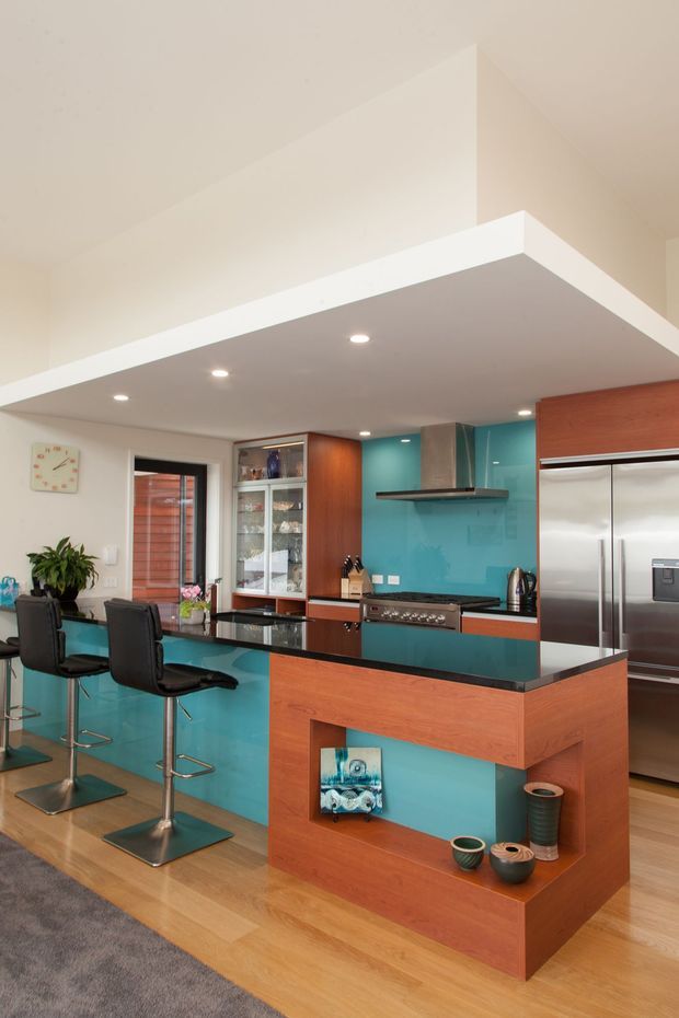 Contemporary open-plan kitchen featuring a large central island with a black polished benchtop, warm timber cabinetry and display shelving, vivid turquoise glass splashback and island panels, stainless-steel appliances, and integrated recessed ceiling lighting. Two bar stools face the island, while a glass-fronted cabinet and timber-lined exterior door are visible in the background. The room has a bright, lively, polished atmosphere with timber flooring and a partial rug in the foreground.