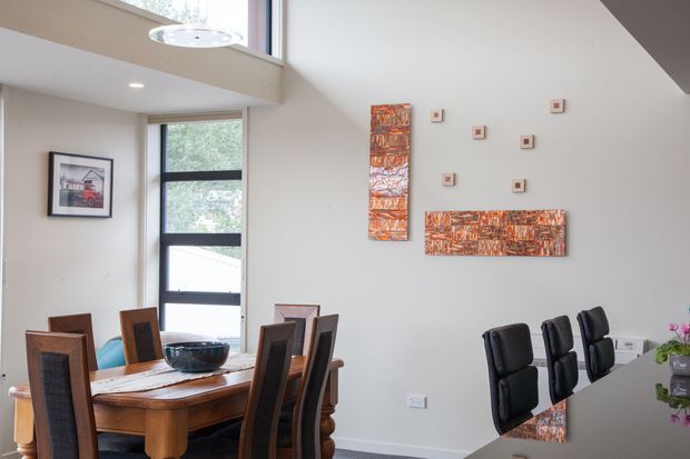 Bright contemporary open-plan dining and kitchen area with a substantial timber dining table and upholstered dining chairs, a glossy dark kitchen island in the foreground, abstract orange-toned wall artworks, and tall windows beneath a high angled ceiling. The room has a partial double-height feel with a modern pendant light and abundant natural light.