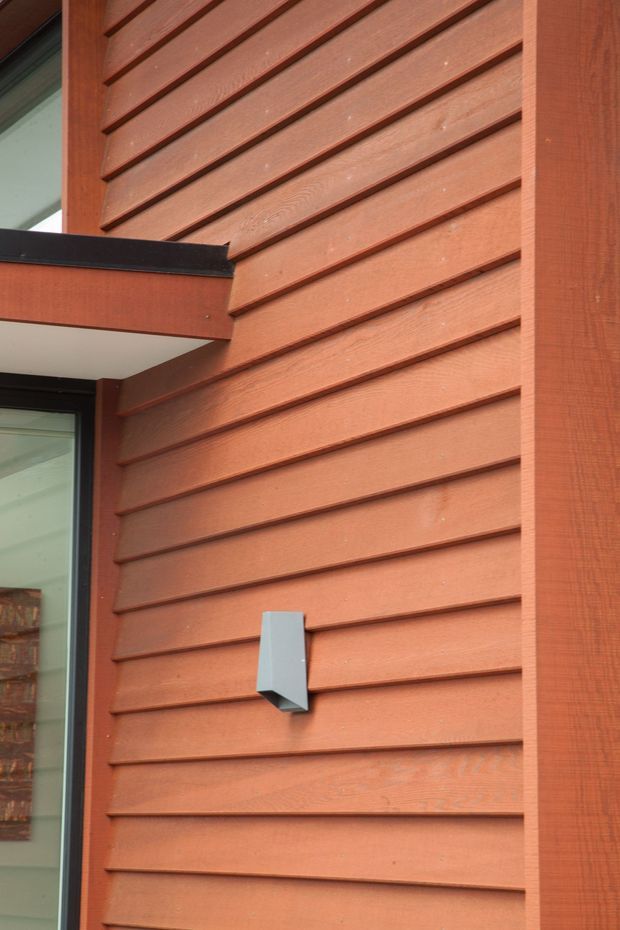 Close-up exterior architectural detail showing reddish-brown horizontal timber weatherboard cladding with pronounced overlapping boards, dark-framed glazing, a projecting trim or balcony edge, and a small modern grey wall light. The image focuses on the building facade and its material detailing.