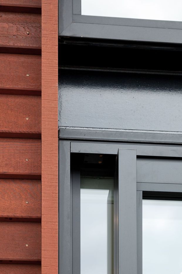 Close-up architectural detail of a dark grey metal-framed window or door set beside reddish-brown horizontal timber weatherboard cladding. The image emphasizes the junction between the textured timber exterior and black/grey painted metal framing, with reflective glazing visible.