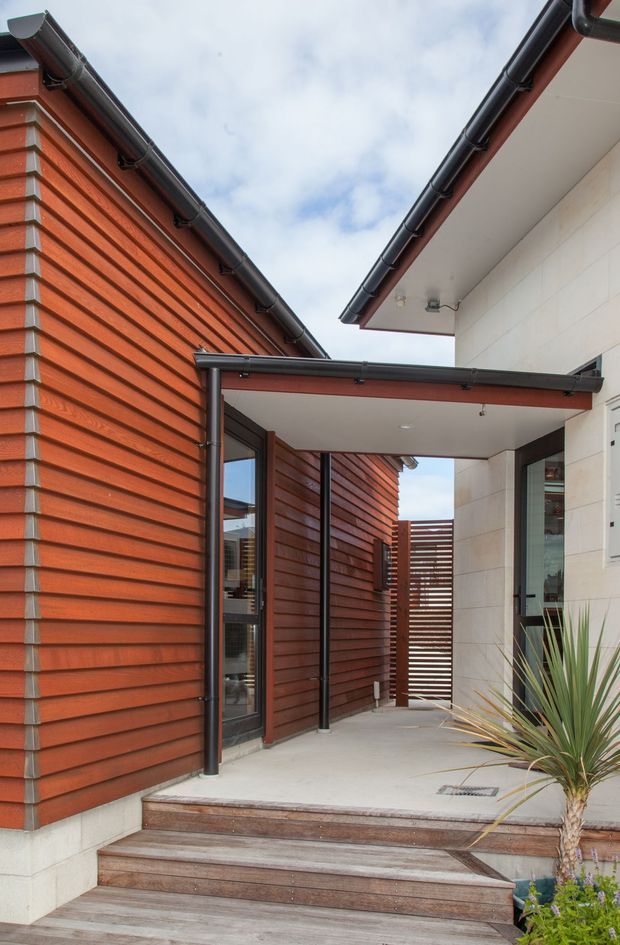 A contemporary residential entry courtyard viewed from outside, with warm horizontal timber cladding, dark metal gutters and downpipes, a covered walkway, pale masonry walls, timber steps and a slatted timber privacy gate. A small planted area sits beside the entrance, and the pitched roof edges frame an open view of the sky.