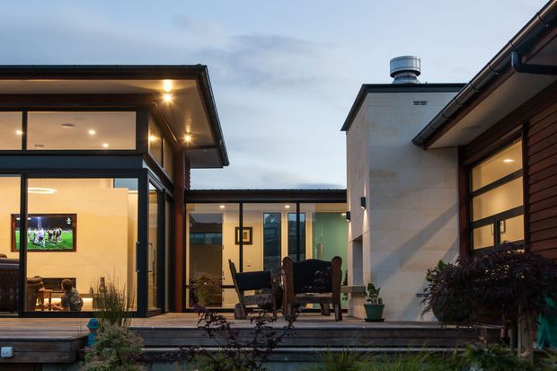 Exterior view of a contemporary courtyard-style residence at dusk, with several connected low-rise wings surrounding a raised timber deck and outdoor seating area. Floor-to-ceiling glazing reveals a warmly lit living space, while dark timber cladding, pale rendered walls, broad roof overhangs, and exposed gutters define the facade.