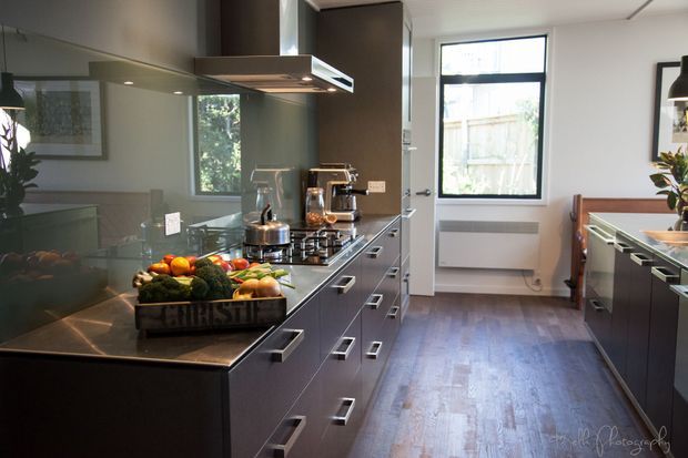 A contemporary galley kitchen with dark grey built-in cabinetry, long stainless-steel benchtops, a reflective glass splashback, integrated appliances, and a central cooking zone with a stainless rangehood. Timber flooring runs through the room, with a large window providing natural light. The space has a streamlined, functional layout with strong dark-and-metallic finishes.