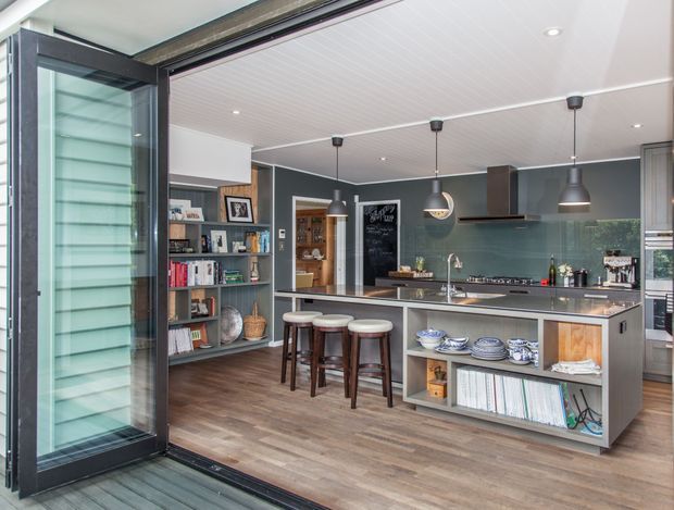 A contemporary open-plan kitchen viewed from an outdoor deck through wide black-framed folding or sliding doors. The room features a large dark island with integrated cabinetry and open shelving, a stainless-steel sink and benchtop, pendant lights, dark cabinetry, timber flooring, and a charcoal feature wall. A built-in shelving unit and stools add functionality and warmth.