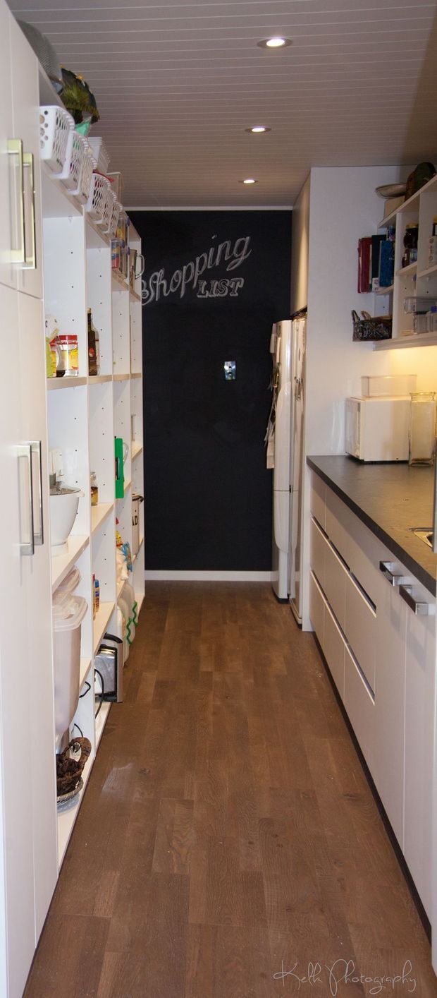 A narrow, well-organised walk-in pantry with tall white open shelving on both sides, integrated white cabinetry and drawers, a dark chalkboard-style feature wall reading “Shopping List,” recessed ceiling lights, and warm brown timber-look flooring. Food containers, baskets, appliances, and pantry supplies are stored throughout the shelving.