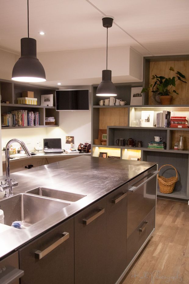 A warm, contemporary kitchen centered on a large island with an extensive stainless-steel benchtop, integrated double sink, and dark built-in cabinetry. Open shelving and display joinery line the rear wall, holding books, crockery, appliances, and decorative items, with a wall-mounted television and two large pendant lights overhead. Timber flooring and warm under-shelf lighting add contrast to the industrial-leaning kitchen fit-out.