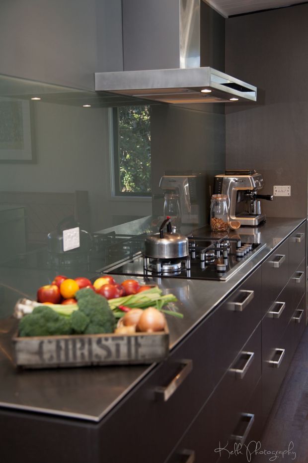 A sleek contemporary kitchen viewed along a long dark island and cabinetry run. The kitchen features a glossy dark benchtop, dark handleless-style drawer fronts with metal pulls, a gas cooktop, stainless-steel kettle, built-in rangehood, espresso machine, reflective glass splashback, and a tray of fresh produce in the foreground. Natural greenery is visible through the window reflection.