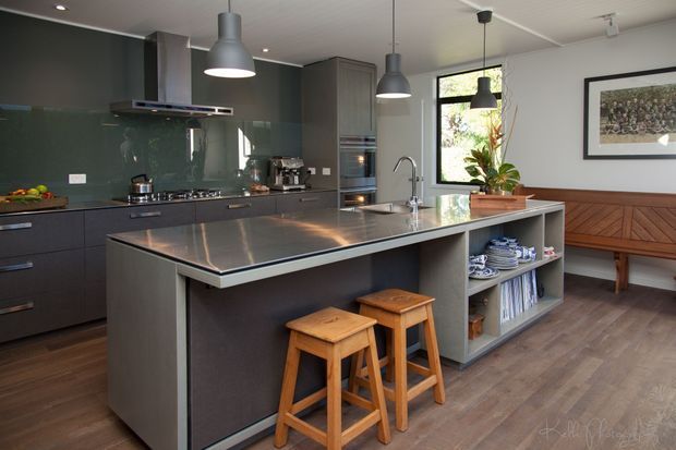 A contemporary kitchen with a large central island, dark cabinetry, stainless-steel benchtop, glossy dark splashback, integrated appliances, pendant lighting, timber bar stools, and an adjacent built-in timber bench seat. The space has warm timber flooring and a framed artwork, creating a refined, practical family kitchen.