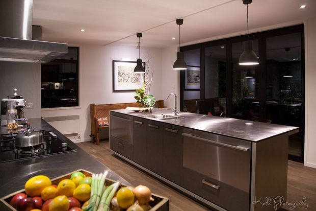 A sophisticated contemporary kitchen featuring a large dark island with an integrated stainless-steel dishwasher and sink, a stainless-steel cooktop and rangehood, dark cabinetry, timber flooring, black-framed glazing, and three pendant lights. A timber bench and dining area are visible beyond the island, with warm low-level lighting and dark exterior views through the glazing.