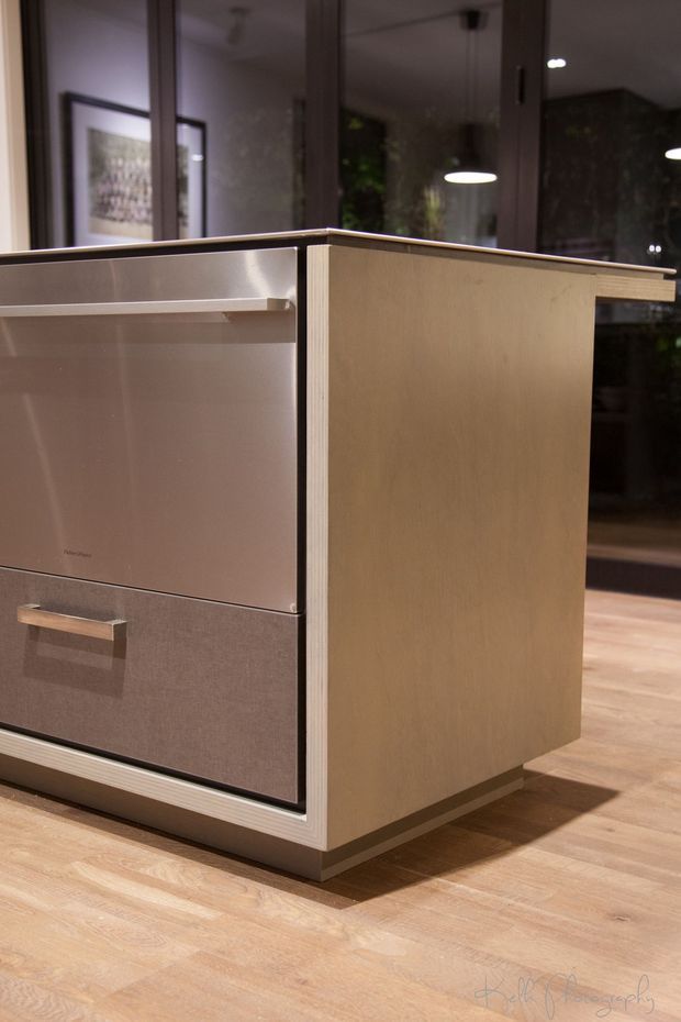 Close-up interior photograph of a compact kitchen island with integrated cabinetry and appliances. The island has pale timber or wood-look panelled sides, a dark contrasting benchtop edge, and flush grey cabinetry with a horizontal metal handle. It sits on a light timber floor in a softly lit contemporary kitchen, with reflective glazing, pendant lights, and indoor greenery in the background.