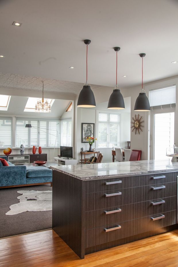Bright open-plan interior centered on a large kitchen island with a polished light stone benchtop and dark wood-grain drawers. Three black pendant lights hang above the island. A living room with a blue sofa, television and rug is visible beyond, along with a dining area, extensive windows with blinds, skylights, and a decorative chandelier. Timber flooring runs through the space.