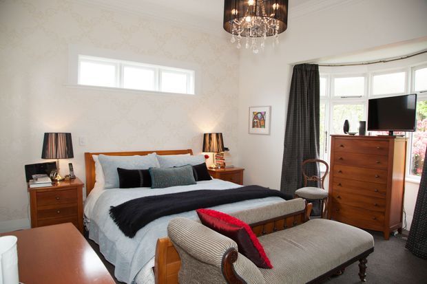 A traditional bedroom with a wooden bed, layered bedding, matching bedside tables and lamps, a tall chest of drawers, upholstered armchairs, patterned wallpaper, drapery, and a decorative chandelier. Bay windows bring in natural light, while dark textiles and warm timber furniture add contrast.