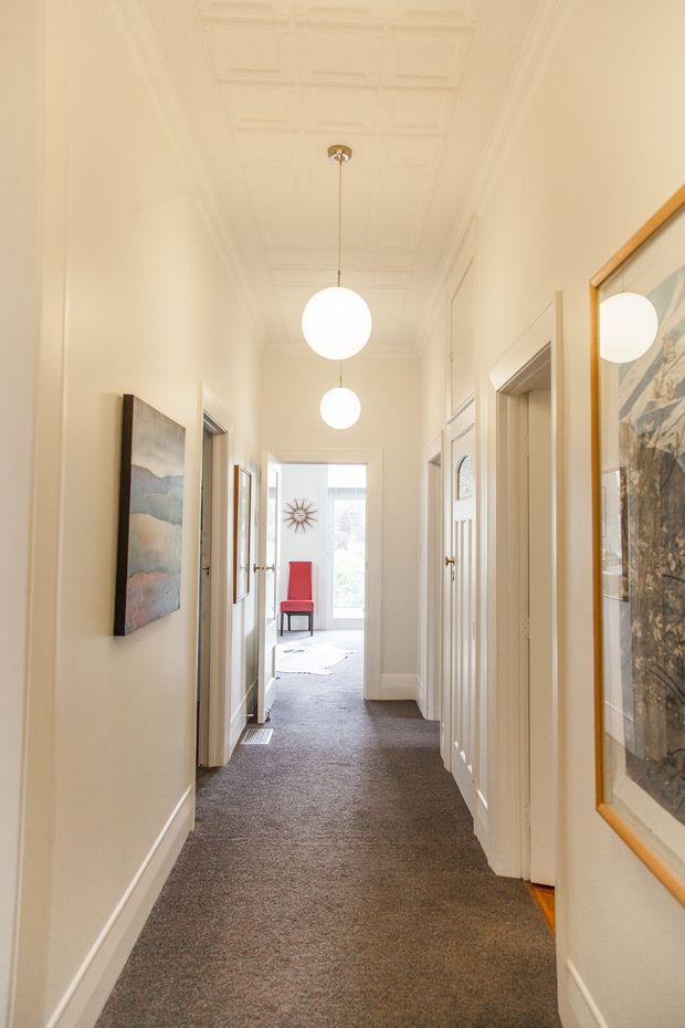 A long, narrow residential hallway with cream-painted walls, decorative white ceiling panelling and mouldings, white trim and multiple doorways. Brown-grey carpet runs through the corridor. Two white globe pendant lights hang centrally, while framed artwork, a mirror, and a red chair are visible toward the brighter room at the end. The space feels bright, refined, and traditionally detailed.