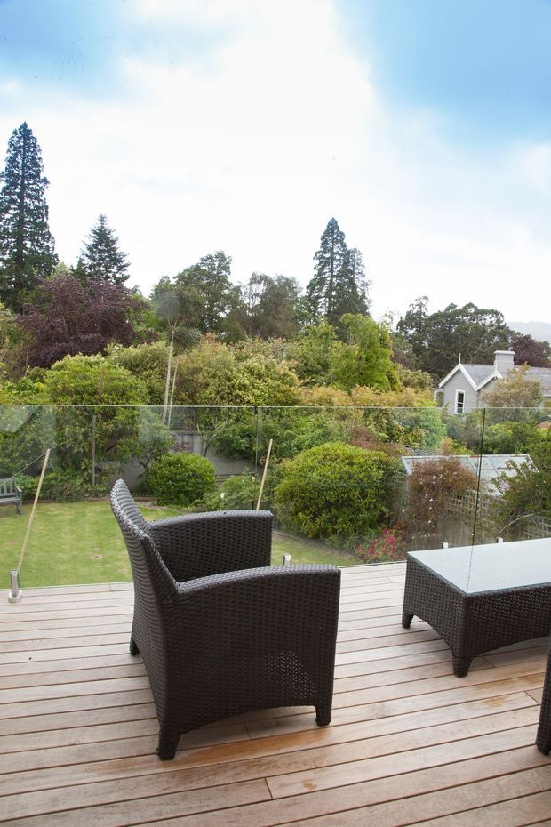 An elevated outdoor timber deck overlooking a lush landscaped garden, with dark woven outdoor armchairs and a matching rectangular table in the foreground. A clear glass balustrade frames the garden view, with mature trees and a neighbouring house visible beyond.