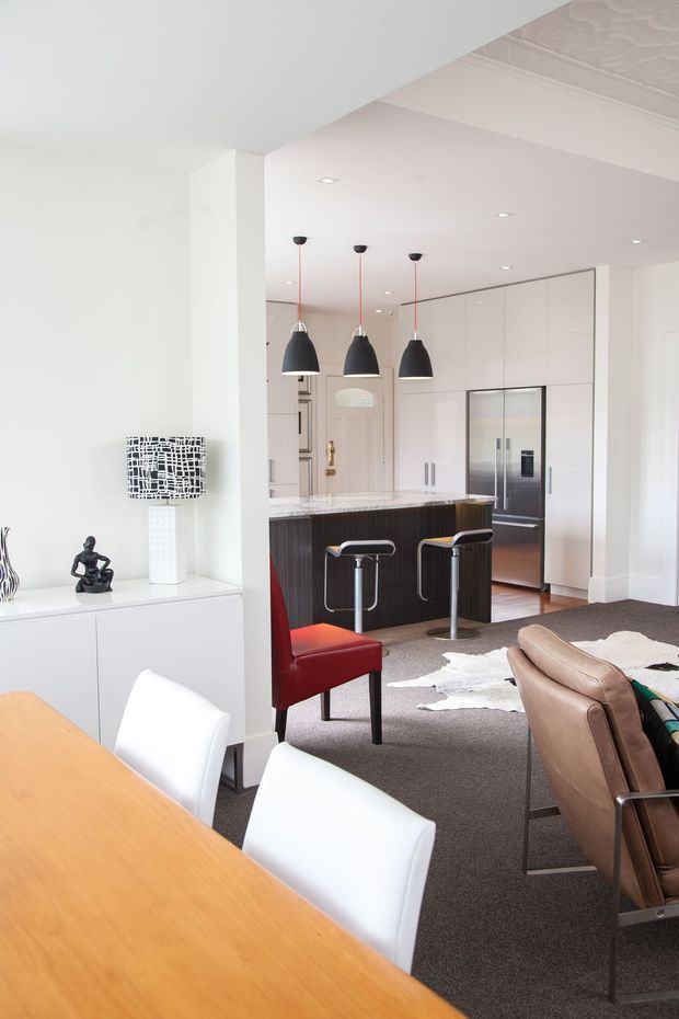 Bright open-plan interior showing a contemporary kitchen connected to a dining and living area. The kitchen features white full-height cabinetry, a stainless-steel refrigerator, a dark wood island with a light stone benchtop, and three black pendant lights. In the foreground is a light timber dining table with white chairs, while the living area includes leather seating, a red accent chair, a white cowhide rug, and minimalist decorative objects.