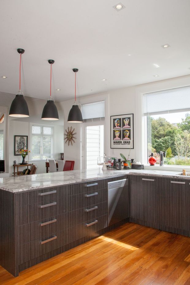 Bright contemporary kitchen with an L-shaped island and peninsula clad in dark vertically grained cabinetry, topped with a light stone benchtop. Three black pendant lights with red cords hang above the island, while recessed ceiling lights and large windows with white blinds provide abundant daylight. Warm timber flooring contrasts with the dark joinery, and a dining area with artwork and decorative objects is visible beyond the kitchen.