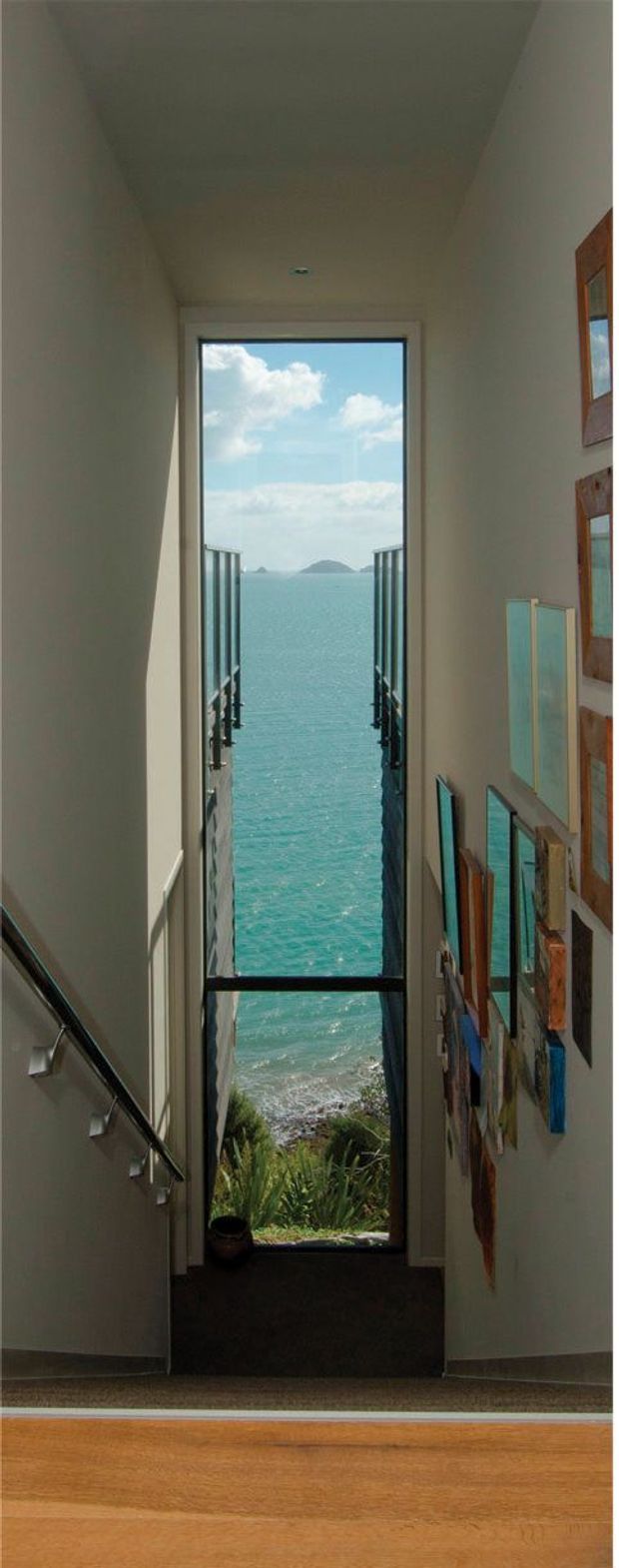 A narrow interior stairway or landing with a tall, floor-to-ceiling glazed opening and dark framed doors overlooking turquoise ocean water, a small island, and coastal vegetation. The space has light painted walls, a recessed ceiling light, dark carpeted steps or landing, warm timber flooring in the foreground, a slim metal stair handrail with small spotlights, and a collection of framed or mounted artwork along the right wall. The view and glazing create a strong vertical coastal focal point.