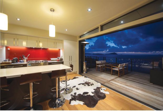 A warmly lit, contemporary open-plan kitchen and dining area opening through large sliding glass doors onto a timber deck. The kitchen features pale cabinetry, a broad light benchtop, a vivid red glass splashback, stainless-steel appliances and a breakfast bar with dark stools. Timber flooring continues toward the outdoor dining setting, with a cowhide rug in the foreground. The deck overlooks a dark waterfront or coastal landscape at dusk.