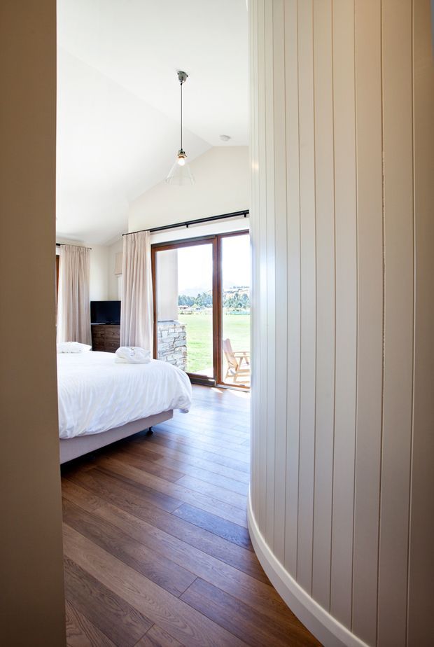 Bright bedroom viewed from an adjoining hallway or entry, featuring a partially visible bed with white bedding, a dark wood dresser and television, pale curtains, a pendant light, and large timber-framed glazed doors opening to a small deck and green landscape. The room has warm timber flooring and a curved white vertical-panel wall or partition in the foreground.