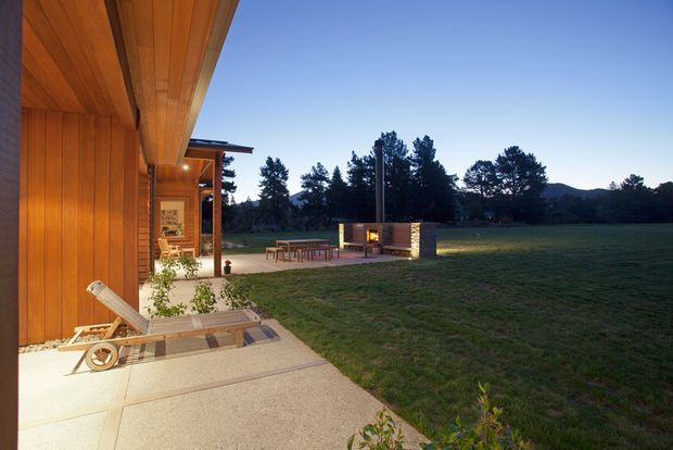 Exterior view of a contemporary timber-clad pavilion or house opening onto a broad lawn. The image shows warm vertical wood cladding and soffit, a concrete terrace with a timber lounge chair, outdoor dining furniture, and a freestanding masonry outdoor fireplace. Dark trees and low hills form the background at dusk.