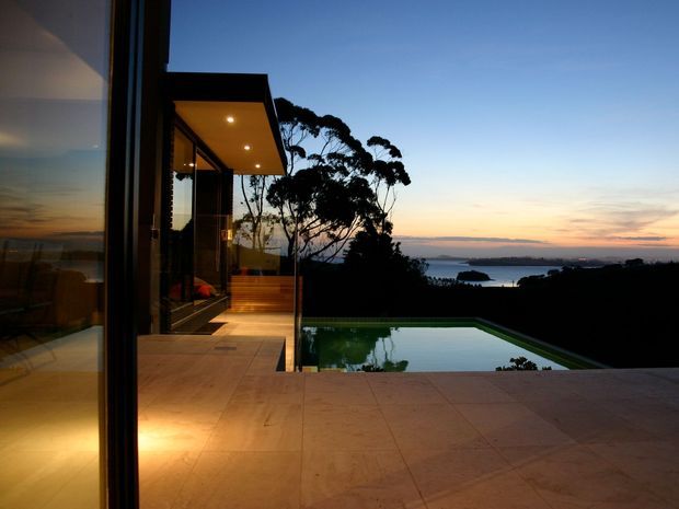 A dusk view from a contemporary house terrace, looking across a tiled pool deck toward an infinity-edge swimming pool and a calm coastal or bayside outlook. Floor-to-ceiling glazing and dark slim frames open onto a covered outdoor area with warm recessed lighting, while the low, rectilinear architecture blends into the surrounding silhouetted landscape.