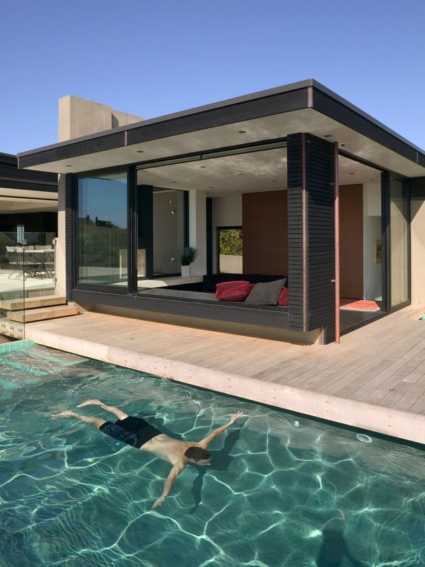 Exterior view of a contemporary flat-roof pavilion beside a swimming pool. The building has extensive floor-to-ceiling glazing, dark horizontal cladding, exposed concrete soffits, and a timber-toned deck. A partially enclosed lounge with cushions is visible inside, while a second terrace and outdoor seating appear to the left.