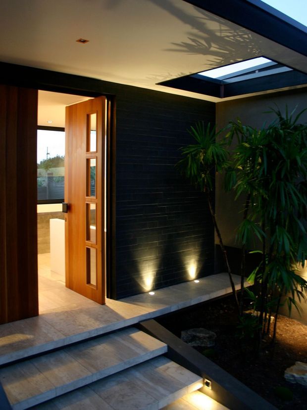 A dramatic covered residential entry courtyard with broad tiled steps leading to a timber pivot-style door with narrow glazed panels. A dark horizontally lined feature wall is illuminated by recessed uplights, while a roof skylight brings daylight into the space and casts plant shadows across the ceiling. Lush potted planting and a small landscaped bed soften the modern, angular entry design.