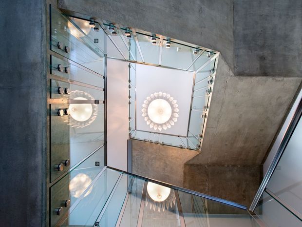 Upward-looking view through a multi-level stairwell or atrium with exposed raw concrete walls, extensive clear glass balustrades, metal framing, and repeated circular pendant lights reflected across the glazing. The image emphasizes the vertical void, transparent circulation edges, and industrial architectural detailing.