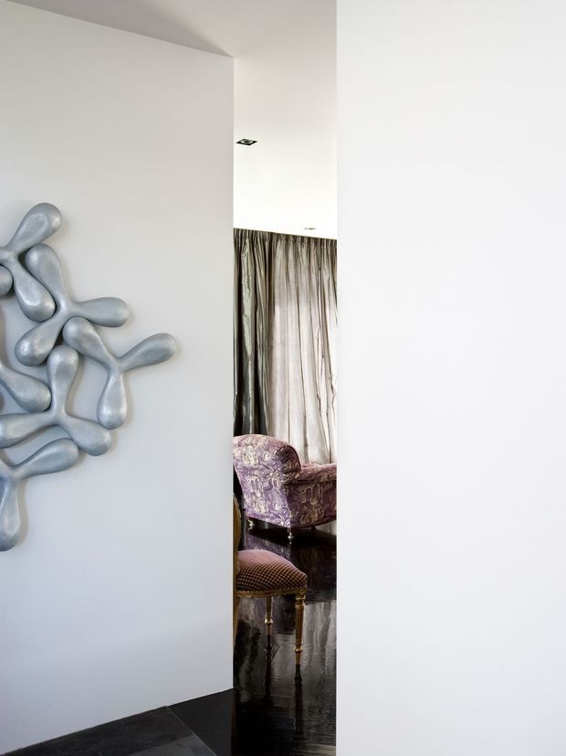 A minimalist interior view through a narrow opening between white walls, revealing an adjoining sitting area with floor-to-ceiling sheer curtains, a patterned upholstered armchair, and a small ornate stool. The foreground features a sculptural organic wall installation in pale grey and dark polished flooring, creating a refined contrast between minimalist architecture and decorative furnishings.