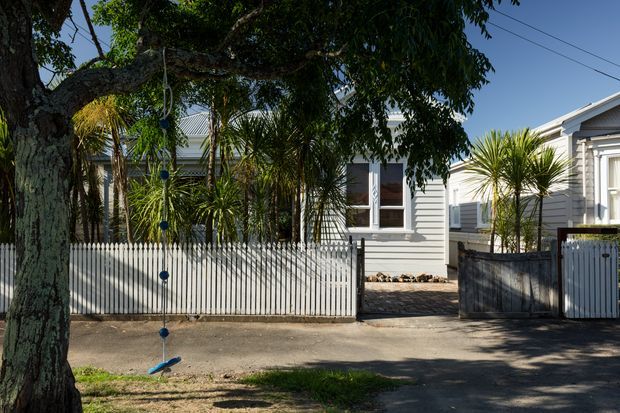 Street-facing view of a compact light-grey weatherboard house behind a white picket fence, with a paved entry path, low front boundary walls, mature trees, and tropical-style planting. The single-storey facade features a pitched metal roof, white-framed windows, and a shaded verandah-like entry area.