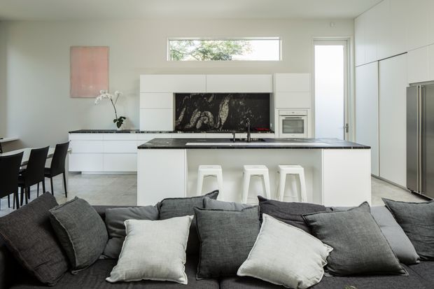 Bright open-plan contemporary kitchen and living area. The kitchen features a large white island with a dark stone benchtop, integrated white cabinetry, a black marble-look splashback, built-in oven, and three white stools. A dining table sits to the left, while a dark grey sectional sofa with contrasting cushions occupies the foreground. White walls, pale flooring, and a narrow horizontal window create a clean, airy interior.