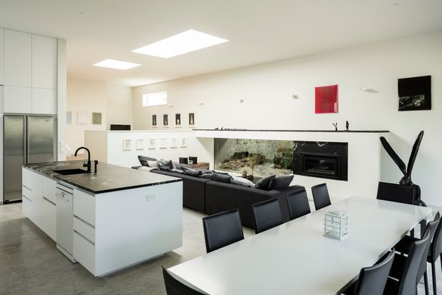 Bright open-plan contemporary interior combining a white kitchen, large island with sink, dining area, and living room. The space features dark stone-like benchtops, white cabinetry, black dining chairs, a dark sectional sofa, recessed linear fireplace, aquarium or glazed feature wall, minimalist artwork, and large rectangular ceiling lights.