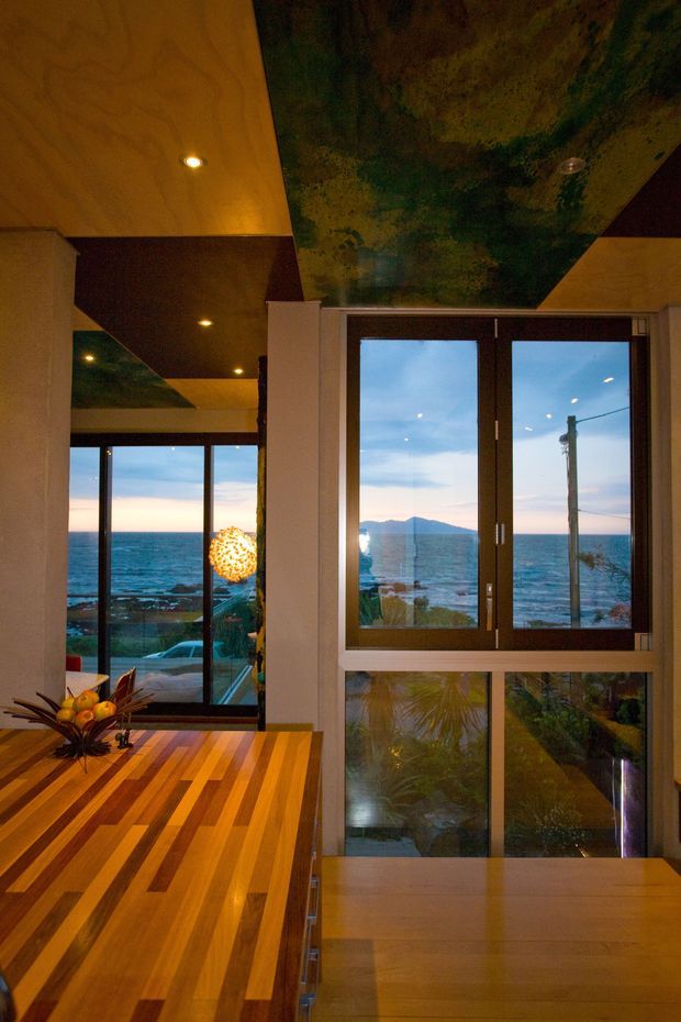A warm, contemporary interior overlooking the ocean, with expansive dark-framed windows and glazed doors. The room features a large striped timber dining table or island in the foreground, warm recessed lighting, pale walls, and a striking mottled green-and-gold feature ceiling. Ocean, coastline, and distant hills are visible through the glazing. A sculptural pendant light and decorative fruit arrangement add warmth.