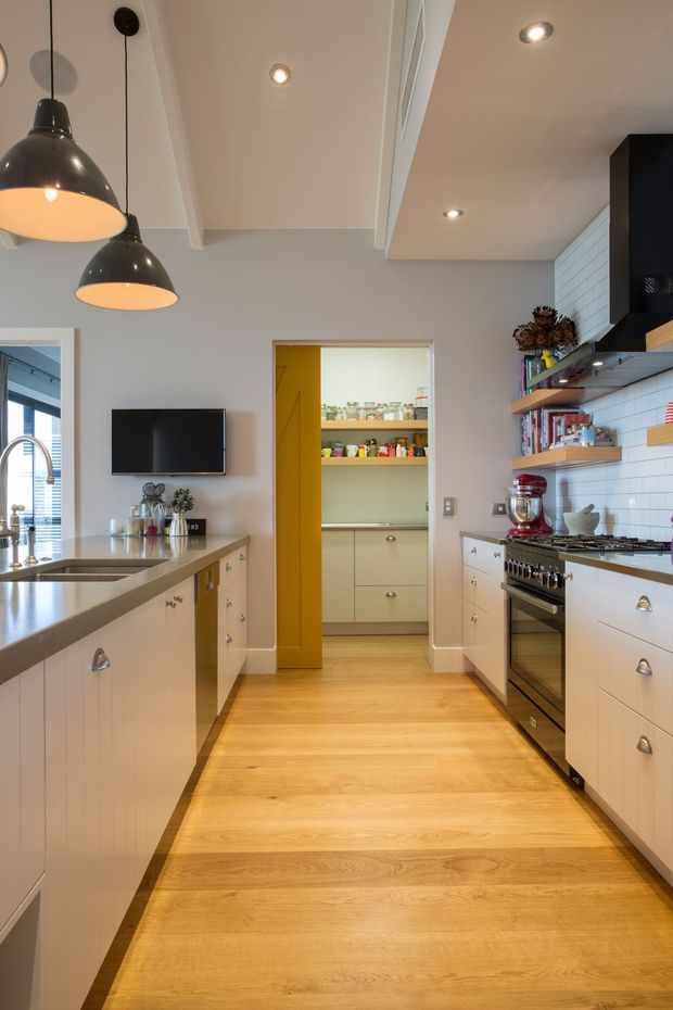A bright, contemporary galley kitchen with a central walkway, pale shaker-style cabinetry, warm timber flooring, white tiled splashback, dark stone-look benchtops, and black-and-chrome appliances. A compact pantry is visible at the rear through a yellow-painted doorway. Open shelving, pendant lights, recessed ceiling lights, and a large island with sink define the space.