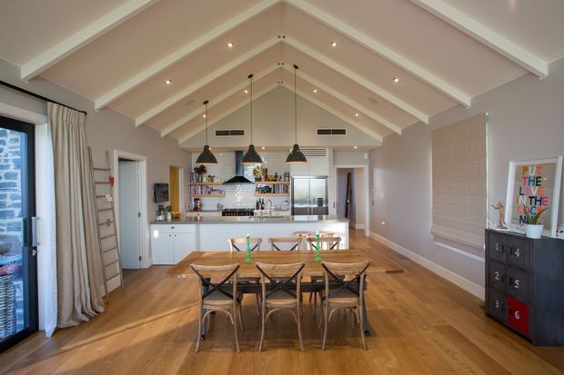 A bright open-plan dining and kitchen interior with a vaulted, exposed-beam ceiling. A timber dining table with six chairs occupies the foreground, while a white kitchen with island, open shelving, patterned backsplash, and pendant lights sits beyond. Warm timber flooring, pale walls, large glazed doors, and soft window coverings create an airy contemporary coastal feel.