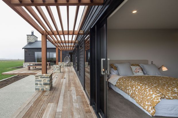A contemporary bedroom opening directly onto a wide timber deck beneath a substantial timber pergola. The room has dark-framed full-height sliding glazing, a grey upholstered bed, patterned mustard-and-white bedding, carpeted flooring, and recessed ceiling lighting. Outside, the deck continues toward a dark-clad pavilion with a chimney, stone-clad low walls, and open rural surroundings.