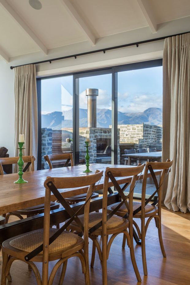 A warm dining room with a long solid timber dining table and multiple timber cross-back chairs. Full-height dark-framed glazing opens the room to a stone-paved outdoor terrace with a built-in stone fireplace and expansive mountain views. The room has a pale exposed-beam ceiling, light neutral walls, timber flooring, and full-length beige curtains on a black curtain rail. Green glass candleholders provide small decorative accents.