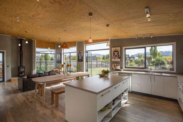 Open-plan kitchen and dining/living space with a large white island, extensive white cabinetry, timber flooring, and a warm plywood-lined ceiling. The room has generous windows and glazed doors overlooking a landscaped suburban setting with hills beyond. A dining table, wood-burning stove, pendant lights, and built-in kitchen storage are visible.