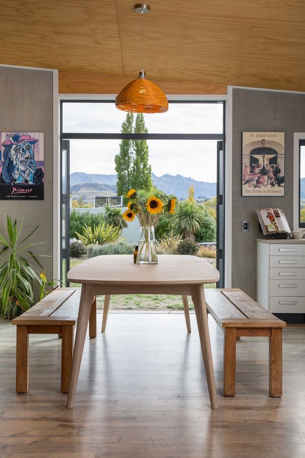 A warm, timber-lined dining area with a solid wood dining table and matching benches positioned in front of large black-framed glazed doors. A woven orange pendant light hangs from the timber ceiling, while grey textured walls, artwork, a sideboard, and indoor plants complete the room. Through the open glazing, a landscaped garden and mountain landscape are visible.