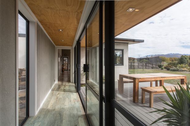 A long, narrow glazed corridor with full-height black-framed sliding glass doors opening onto a timber deck and outdoor dining area. The corridor has a warm plywood-lined ceiling, pale textured walls, and a light stone or concrete-look floor. The view extends toward a contemporary single-storey pavilion and open landscaped surroundings.