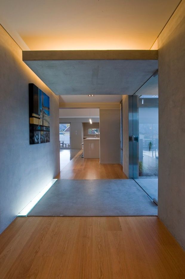 A modern residential hallway viewed toward an open-plan kitchen and living area. The corridor features warm timber flooring, smooth grey concrete or plaster surfaces, a large exposed concrete overhead element, concealed linear lighting, and full-height glazing on the right. The composition has clean geometric lines and a strong indoor-outdoor connection.