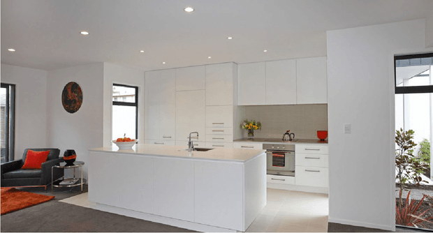 Bright contemporary open-plan kitchen with extensive white flat-panel cabinetry, a large white island with integrated sink, pale tiled splashback, and stainless-steel appliances. The kitchen opens to a living area with dark grey flooring and red-orange accents, while black-framed glazing brings in natural light.