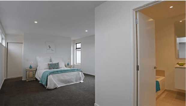 A bright contemporary bedroom with a neatly made bed, turquoise and white bedding, dark carpet, white walls, recessed ceiling lights, and narrow vertical windows. An open doorway reveals an adjoining bathroom with a vanity and freestanding or partially visible bathtub.