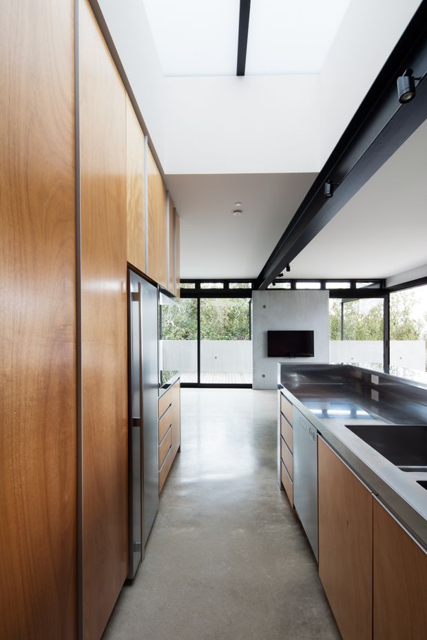 A contemporary open-plan kitchen viewed along a narrow circulation aisle. Full-height warm timber cabinetry lines the left wall, while a large island with dark stone or stainless-steel benchtops and integrated sink occupies the right. Polished concrete flooring, black-framed floor-to-ceiling glazing, exposed dark structural beams, and a bright skylight create a clean, industrial-modern interior. A concrete media wall with a television is visible beyond the kitchen.