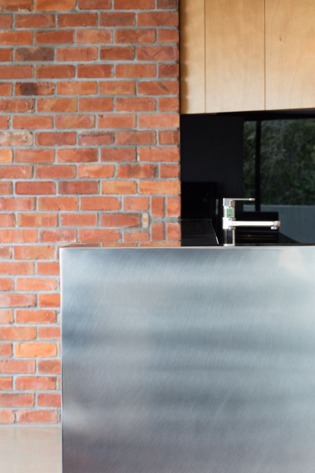 Close-up architectural detail of a contemporary kitchen, featuring a brushed stainless-steel island or benchtop, exposed red brick wall, black splashback and cabinetry, light timber overhead cabinetry, and a chrome mixer tap. The image focuses on the material contrast between brick, metal, timber, and black finishes.