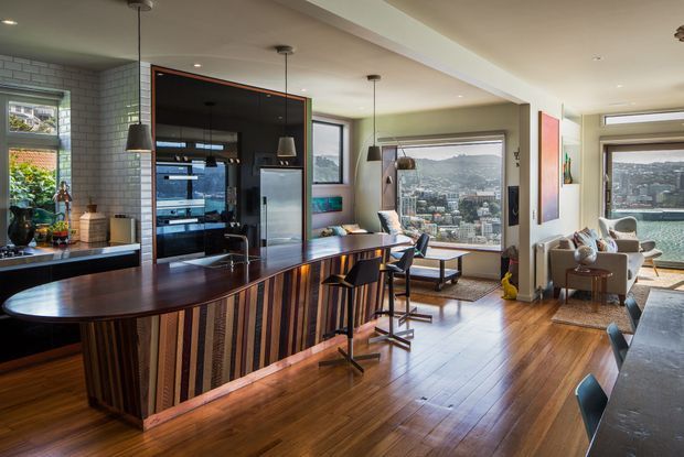 Open-plan contemporary kitchen and living area with a large curved island featuring vertically striped timber panelling, dark stone benchtop, integrated sink and bar seating. The kitchen includes dark cabinetry, a tiled splashback and pendant lighting, opening to a timber-floored dining and lounge space. Large windows provide expansive urban and water views.